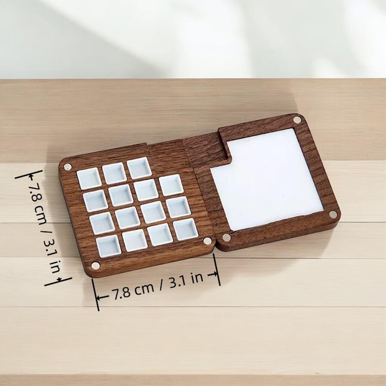 May include: A wooden paint palette with 16 white paint wells. The palette is 7.8 inches wide and 7.8 inches long. It has a hinged lid with a blank white surface.