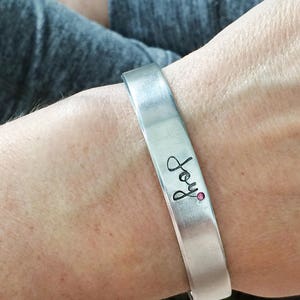 May include: A silver bracelet with the word "Joy" engraved on it. A small pink stone is set in the metal.