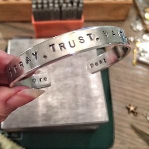 May include: Silver bracelet with the words "Pray. Trust. Wait." engraved on it. The bracelet is on a wooden surface.