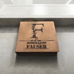Puede incluir: Un álbum de fotos de cuero marrón con una inscripción personalizada. La inscripción dice "EST. 06/06/2024 JENNIFER & KURT FAUSER" y presenta una letra "F" estilizada con un diseño floral.