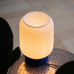 May include: A white ceramic table lamp with a blue base and a ribbed shade. The lamp is turned on and emitting a warm glow.