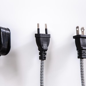 Pode incluir: Tr&ecirc;s tipos diferentes de tomadas el&eacute;tricas com cabos pretos e cabos com padr&otilde;es brancos e pretos. As tomadas est&atilde;o sobre um fundo branco.