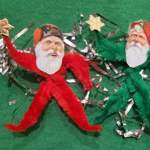May include: Three vintage Christmas ornaments shaped like Santa Claus figures. Each Santa has a painted face, a white beard, and a star in one hand. The figures are red, green, and white, with silver tinsel and candy cane accents.