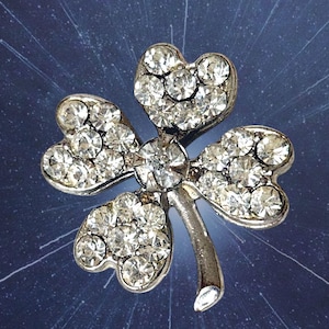 May include: A silver-toned four-leaf clover brooch featuring a metallic frame encrusted with numerous clear, sparkling rhinestones. The design includes a central stone and a curved stem extending from the base of the clover leaves.