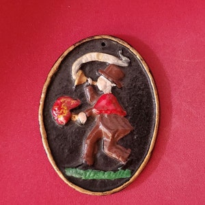 May include: An oval-shaped, black cast iron ornament with a gold rim. The ornament depicts a person in a red and brown costume playing a horn. The person is standing on a green patch of grass.
