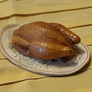 May include: A miniature brown roasted chicken figurine resting on a small, oval-shaped ceramic platter. The platter features a cream-colored base with a delicate, repeating pattern of small blue flowers and green leaves along its outer edge.