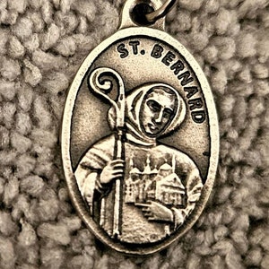 Könnte beinhalten: Ovale Silberanhänger mit einem Bild von St. Bernhard, der einen Stab und ein Gebäude hält. Der Text "ST. BERNARD" ist über dem Bild gewölbt. Der Anhänger hat oben eine kleine Schlaufe für eine Kette.