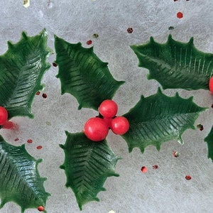 May include: Three sprigs of artificial holly with green leaves and red berries on a white background with red, gold, and silver glitter.