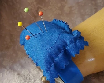 Può includere: Un cuscino per spilli in tessuto blu con un elastico blu attaccato a una mano di legno. Il cuscino per spilli ha quattro spilli colorati inseriti al suo interno.