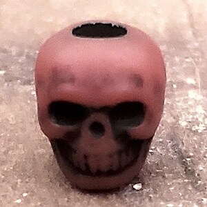 May include: A small, matte, dark red skull-shaped bead with a smooth finish. The bead features a central hole at the top, deep-set eye sockets, a nasal cavity, and a detailed jawline with visible teeth, all rendered in a solid, uniform color.