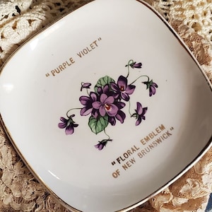 May include: Square white plate with gold trim featuring a purple violet floral design and the text "PURPLE VIOLET" and "FLORAL EMBLEM OF NEW BRUNSWICK". The plate is resting on a cream-colored lace doily.