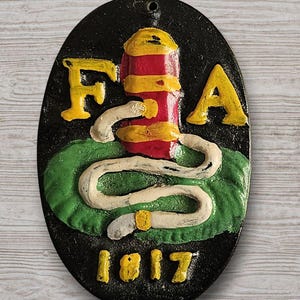 May include: Oval-shaped, black plaque with a relief design. Features the letters "F A" in yellow, a red and yellow column, and a white snake on a green base. The number "1817" is at the bottom. A hole is at the top for hanging.