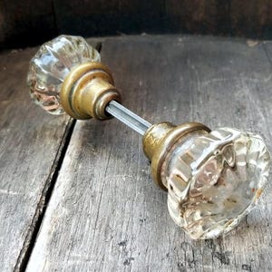May include: Vintage glass door knob with brass fittings. The clear glass knobs feature a cut design, and the brass has a warm, aged patina. The door knob is mounted on a metal rod and rests on a weathered wooden surface.