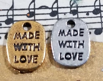 May include: Two oval-shaped metal charms, one in a gold tone and one in a silver tone, each featuring a small hole at the top for attachment. The text MADE WITH LOVE is embossed in black capital letters on the front surface of both metal charms.