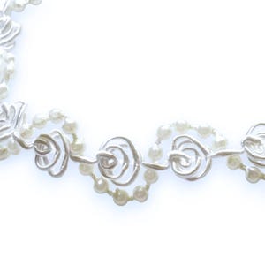 May include: A delicate necklace featuring a repeating pattern of white pearl beads and silver-toned floral accents. The design incorporates small, round pearls and stylized rose-shaped metalwork, creating an elegant and feminine aesthetic.