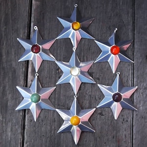 May include: Seven silver star ornaments with colorful center gems, including red, yellow, green, and purple. Each star has a loop for hanging and a pink accent. The ornaments are arranged on a weathered wooden surface.