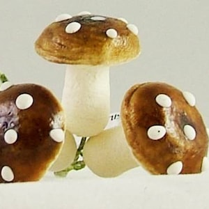 May include: A collection of decorative mushroom figurines. The mushrooms have brown caps with white spots and off-white stems. One mushroom stands upright, while two others are positioned on their sides. The background is a plain white.