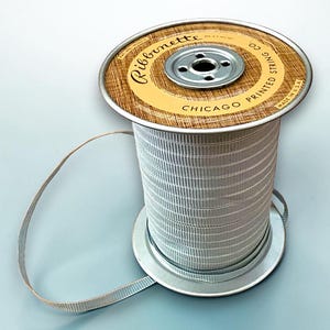 May include: A spool of light gray ribbon with a ribbed texture, wound around a metal core. The top label is tan with a wood grain pattern, featuring the text Ribbonette, Chicago Printed String Co, and Made in U.S.A. in black lettering.
