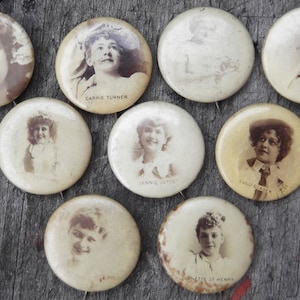 May include: A collection of ten vintage pinback buttons featuring sepia-toned portraits of women. Some buttons have names like "Carrie Turner", "Jennie Hyde", and "Lulu Glaser" printed on them. The buttons are arranged on a weathered wooden surface.