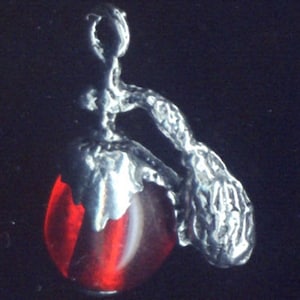 Puede incluir: Colgante de plata con una gema roja de forma ovalada. El colgante tiene un diseño abstracto y orgánico con una superficie texturizada. La gema es translúcida, con una raya blanca en el centro.