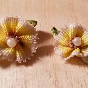 Pode incluir: Dois acessórios de cabelo de flores feitos à mão. Cada flor é amarela e branca com um centro de pérola e folhas de fita verde. Os acessórios estão em uma superfície de madeira clara.