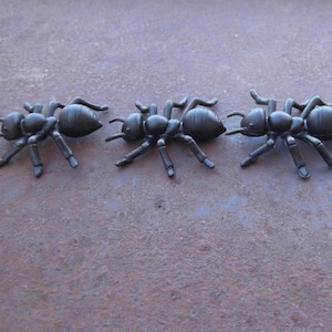 May include: A row of black plastic ant figurines. The ants are detailed with six legs, segmented bodies, and antennae. They are arranged in a line on a textured, brown surface, suggesting a decorative or educational theme.