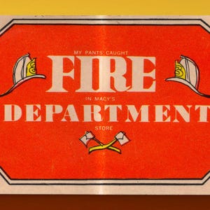 May include: A rectangular orange sign with a black border featuring the text MY PANTS CAUGHT FIRE IN MACY'S DEPARTMENT STORE. The design includes two white firefighter helmets with yellow accents on the sides and two crossed yellow axes below.