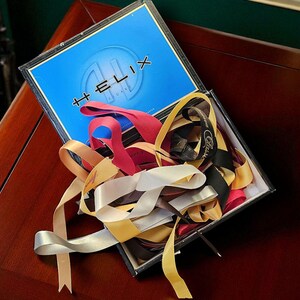 May include: An open box with a blue lid featuring the word "HELIX" and a variety of colorful ribbons. Ribbons in shades of red, gold, black, and silver spill out of the box, creating a visually appealing display.