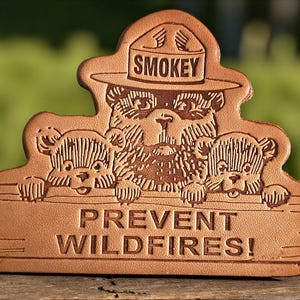 以下が含まれることがあります： スモーキーベアと2匹の子熊が描かれた茶色の革製キーホルダー。「SMOKEY」の文字が帽子に、「PREVENT WILDFIRES!」の文字が木製の看板に書かれています。デザインは革にエンボス加工されています。