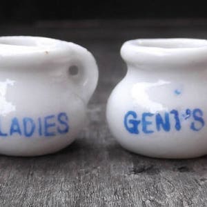 May include: Two white ceramic miniature toilets with blue lettering. One says "LADIES" and the other says "GENT'S".