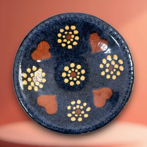 Puede incluir: Un plato redondo de cerámica con un esmalte azul oscuro moteado. La superficie presenta una flor central de puntos amarillos, rodeada por tres flores adicionales de puntos amarillos y tres motivos marrones en forma de corazón en patrón alterno.