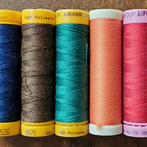 May include: Five spools of thread in different colors: dark blue, brown, teal, orange, and pink. The spools are labeled with the brand name "Amann" and "ASPO" and the thread type "100% Polyester".