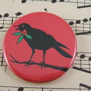 May include: A red button with a black silhouette of a bird holding a sprig of holly with red berries. The bird is perched on a branch.