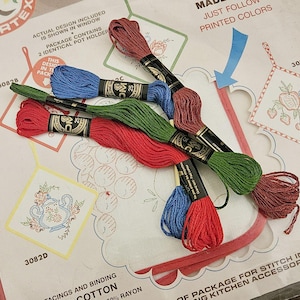 Op de afbeelding: Een close-up van een naaldpuntset met vijf spoelen borduurgaren in rood, groen, blauw, roest en bruin. De set bevat twee identieke theedoeken met een bedrukt ontwerp van druiven en een theepot. De set bevat ook instructies voor het borduren en keukengerei. De set is gelabeld met "Colortex" en "3082A".
