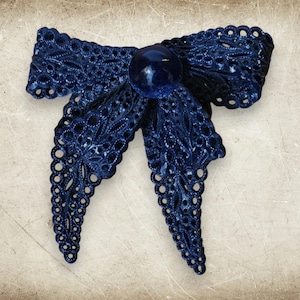 May include: A dark navy blue bow-shaped brooch featuring an intricate, openwork lace-like pattern throughout its structure. A smooth, round, dark navy blue bead is centered at the knot of the bow, providing a solid contrast to the detailed texture.