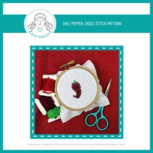Puede incluir: Un patrón de punto de cruz de un chile rojo sobre tela Aida blanca en un aro de bordado de madera. El aro está sobre una superficie de tela roja con hilo de bordar rojo, burdeos y verde.