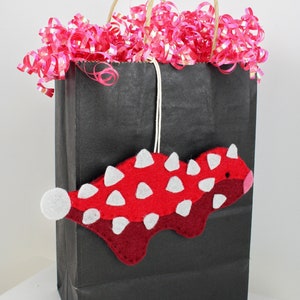 May include: A black paper gift bag with pink and silver curling ribbon. A red and white felt dinosaur ornament hangs from the bag.