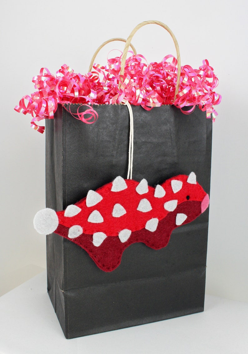 May include: A black paper gift bag with pink and silver curling ribbon. A red and white felt dinosaur ornament hangs from the bag.