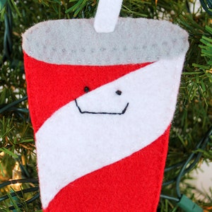 May include: A red and white felt Christmas ornament shaped like a soda can with a straw. The can has a smiling face.