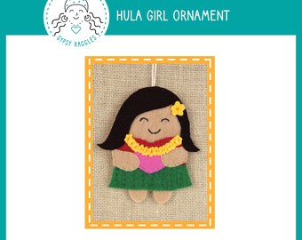 Puede incluir: Un adorno de fieltro de una bailarina de hula con un collar de flores amarillo, un vestido rosa y una flor amarilla en el pelo. El adorno está sobre un fondo de arpillera.