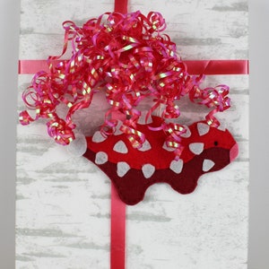 May include: A white gift box with a red ribbon and a red and white felt animal ornament. The box is wrapped in white paper with a wood grain pattern.