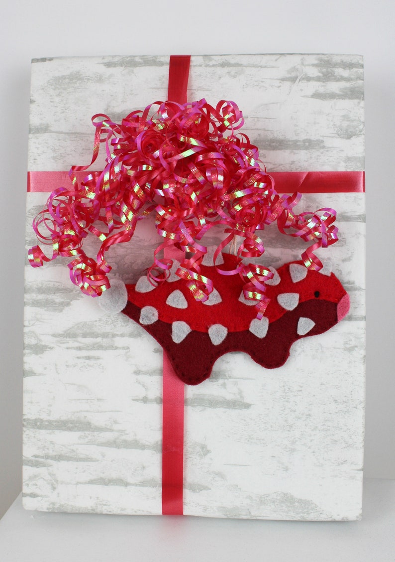 May include: A white gift box with a red ribbon and a red and white felt animal ornament. The box is wrapped in white paper with a wood grain pattern.