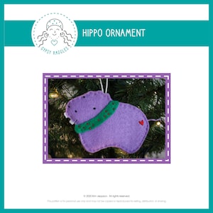 Puede incluir: Adorno de hipopótamo de fieltro morado con una bufanda de fieltro verde y un detalle de corazón de fieltro rojo, colgado de un árbol de Navidad.