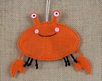 May include: Orange felt crab ornament with black eyes, a smiling mouth, and black legs. The crab is hanging from a white string.