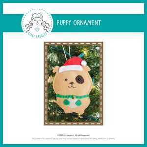 Può includere: Un ornamento a forma di cucciolo in feltro con cappello di Babbo Natale, corpo marrone e una macchia nera. L'ornamento, con un collare verde, è appeso a un albero di Natale. Il testo "PUPPY ORNAMENT" è visualizzato sopra.