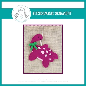 May include: A pink felt plesiosaurus ornament with white polka dots and a green bow. The ornament is hanging from a string and has a happy face.