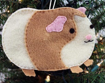 May include: A felt Christmas ornament shaped like a guinea pig. The ornament is white with brown and pink accents. The guinea pig has a pink ear, brown fur, and black eyes.