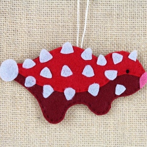 May include: Red and maroon felt ornament shaped like a dinosaur with white felt spikes. The ornament has a white string for hanging.