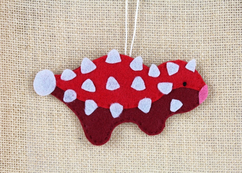 May include: Red and maroon felt ornament shaped like a dinosaur with white felt spikes. The ornament has a white string for hanging.