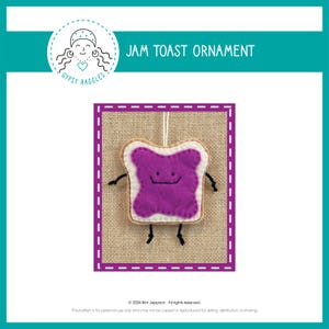 Może przedstawiać: Filcowa ozdoba w kształcie kromki chleba tostowego z fioletowym dżemem i uśmiechniętą buzią. Ozdoba jest zawieszona na sznurku i ma brązowe płócienne tło. Tekst "JAM TOAST ORNAMENT" znajduje się w górnej części obrazu.